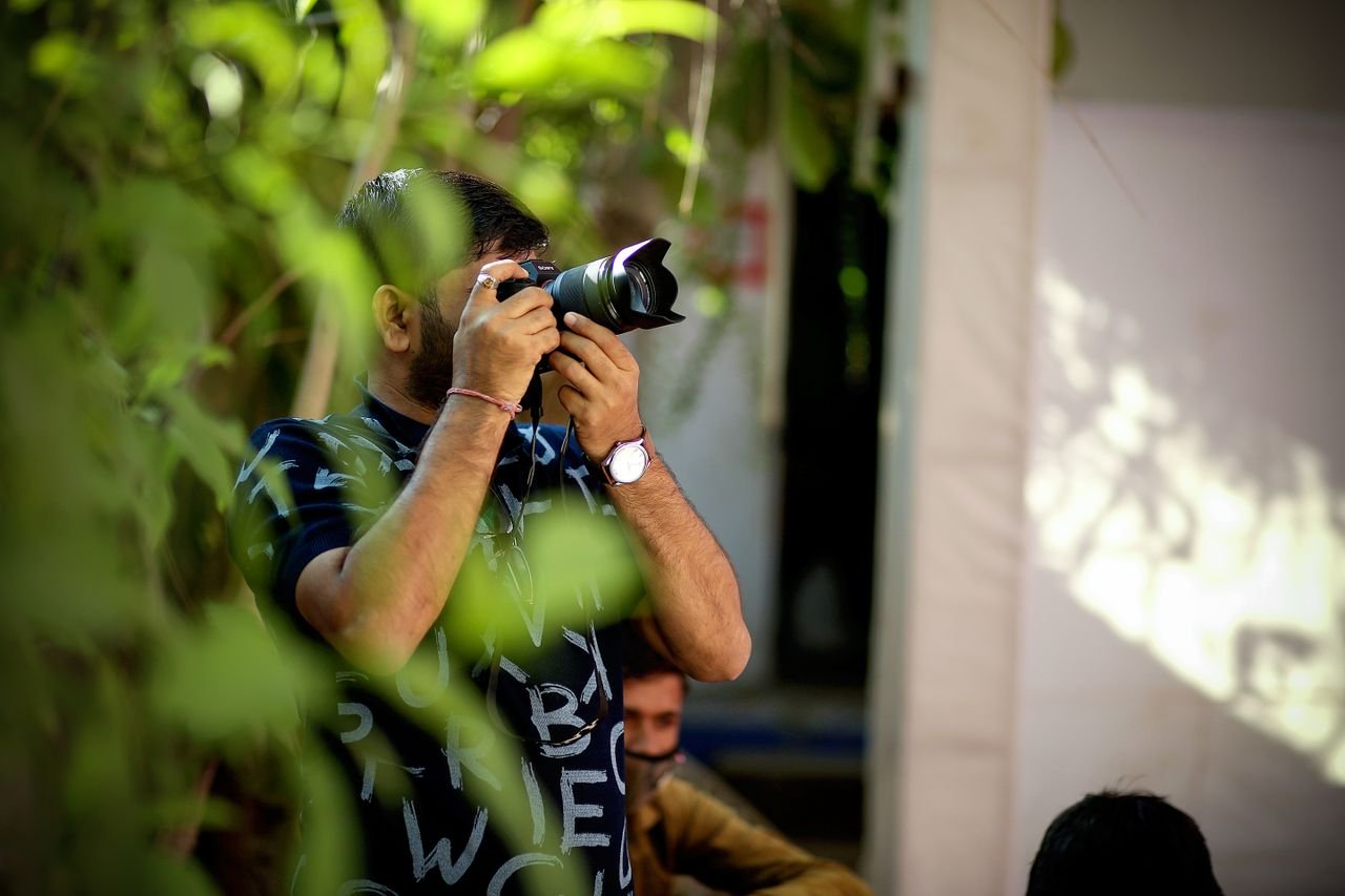 Male photographer taking pictures outdoors with a DSLR camera, surrounded by greenery.